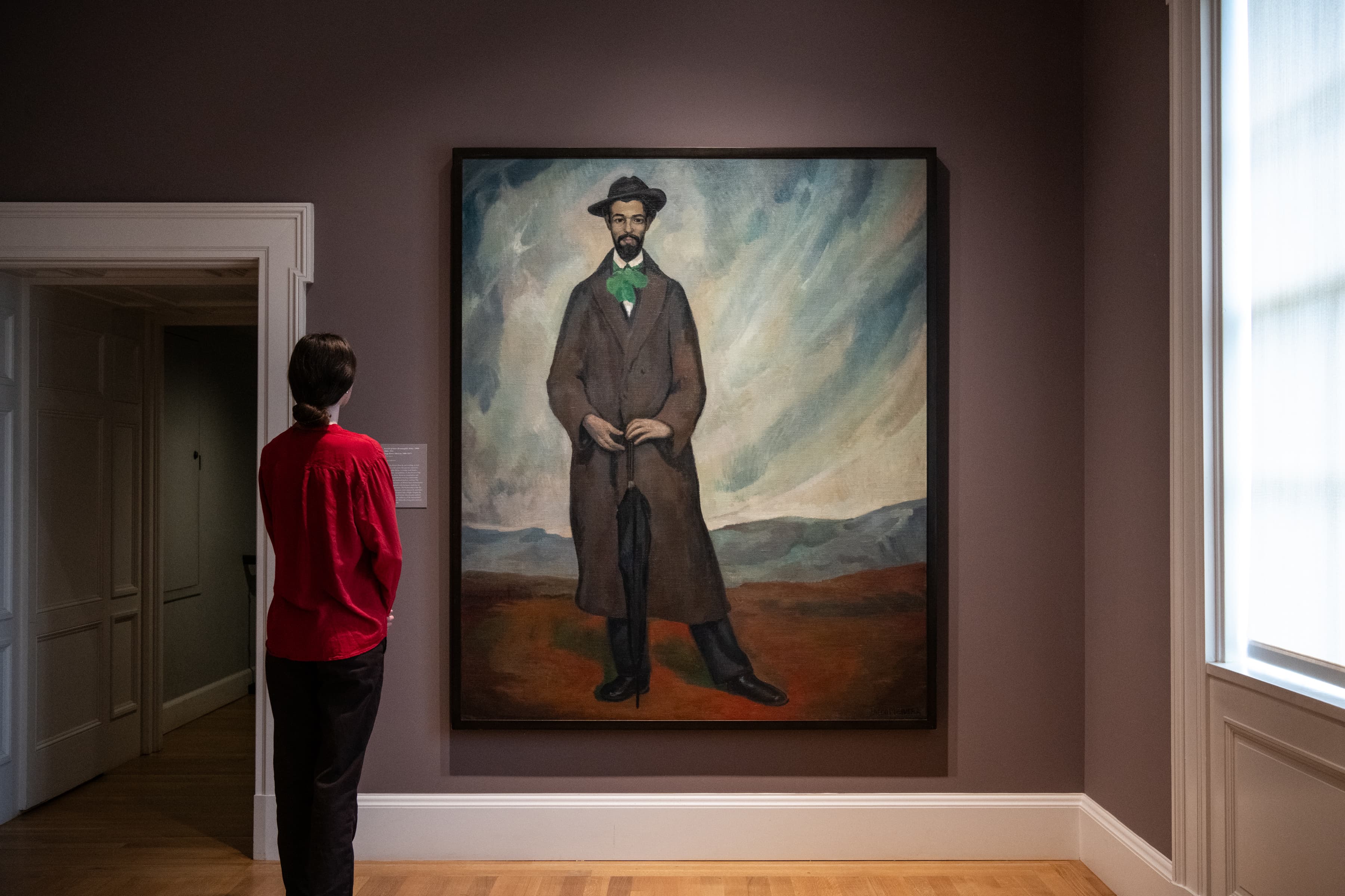 A person looks at a painting of a man wearing a sophisticated gentleman wearing a fashionable overcoat, green feathered necktie, and hat. while standing on an open plain with mountains in the background.