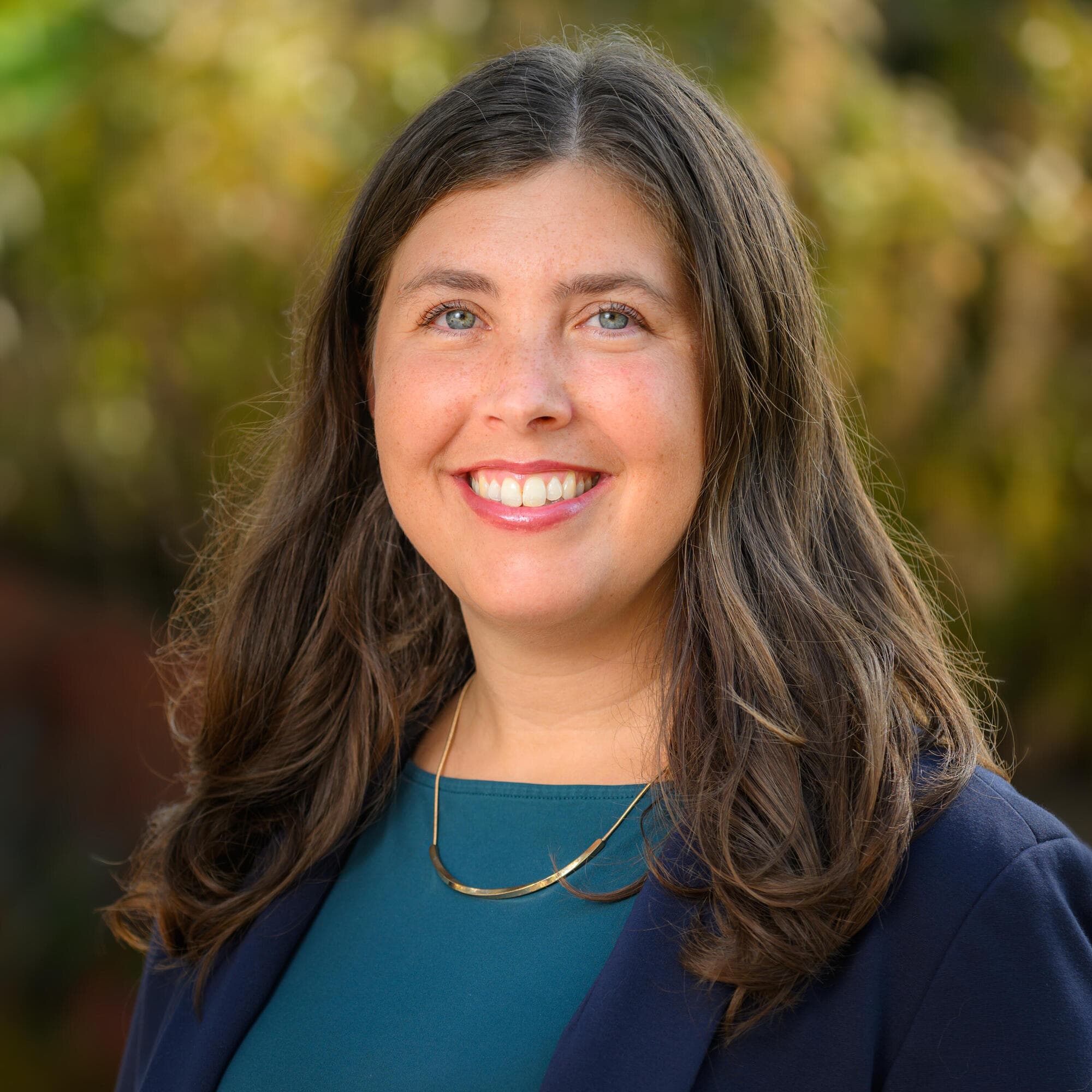 Person with long hair in a dark blue blazer and teal top smiling outdoors.