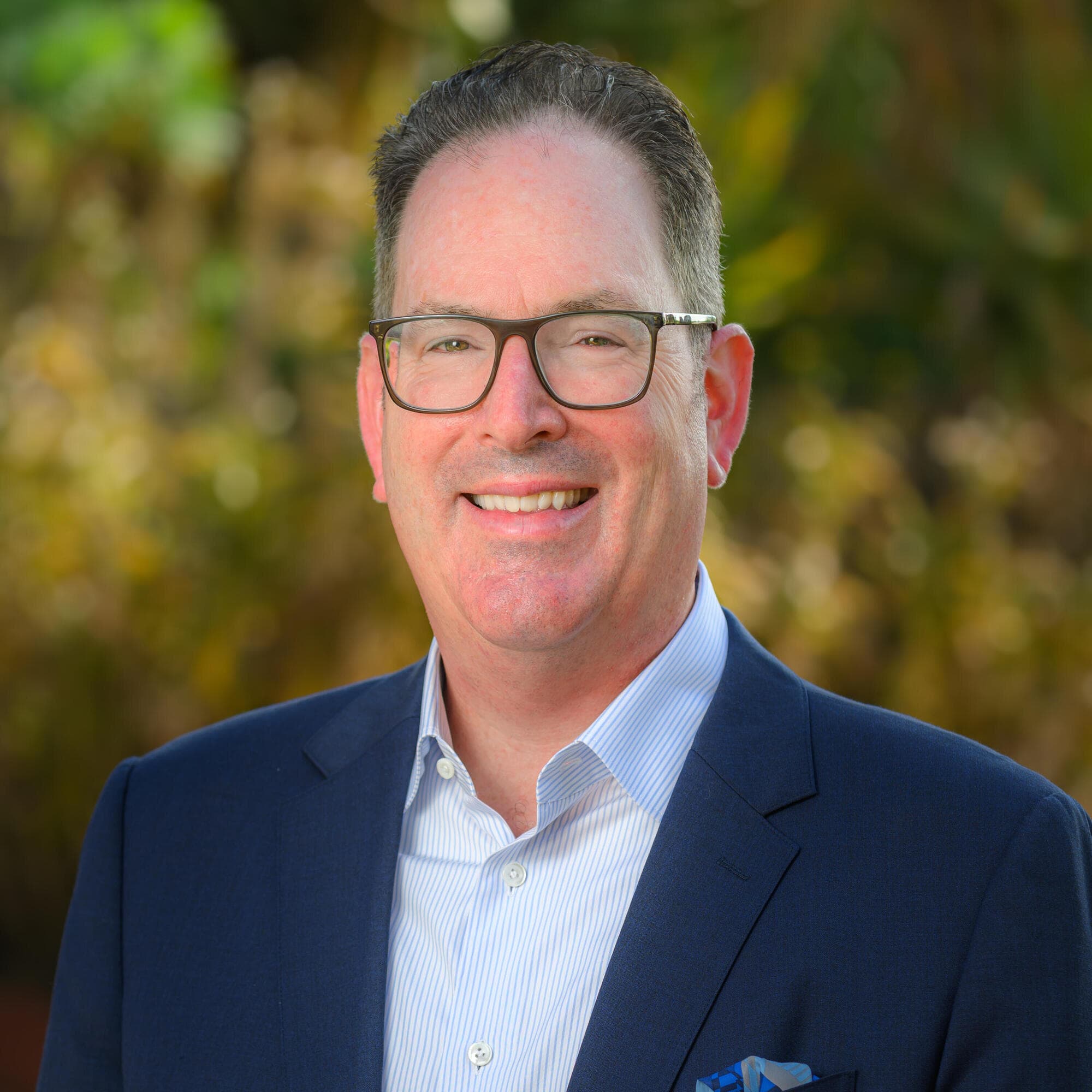 Person with glasses in a dark suit jacket smiling outdoors.