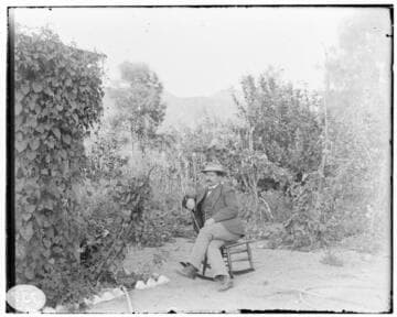 A man sitting in a rocking chair in the garden of a residence.  same shot as 01-