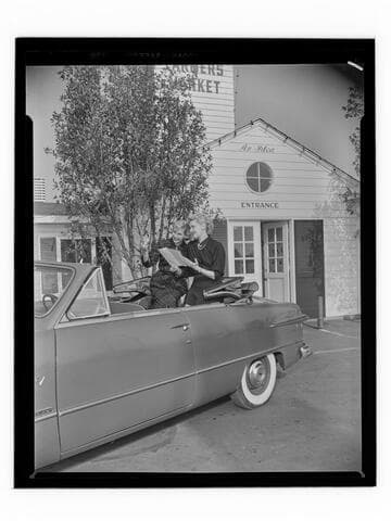 Two women posed in a car in front of the Farmers Market, Los Angeles