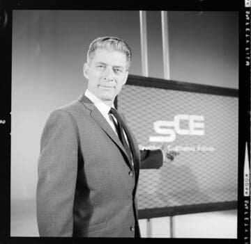 Announcer on TV-Film set speaking to camera with SCE logo over his shoulder