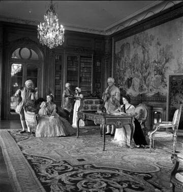 Six people in period costume in the west end of the library of the Huntington residence